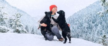 A youthful woman, bundled up in cozy attire, strolls through a scenic snowy mountain landscape, accompanied by her two dogs.